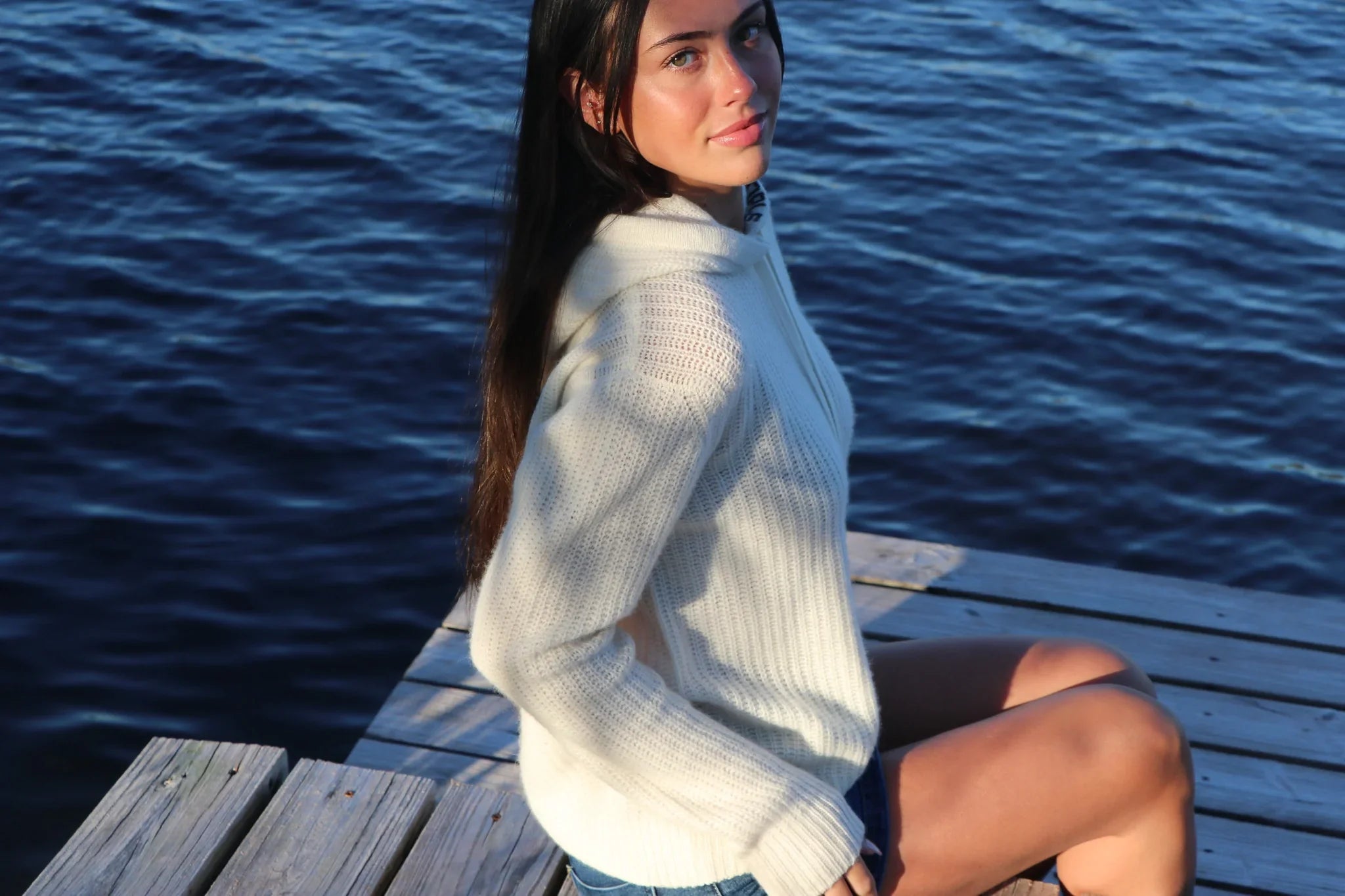 Woman in a white cashmere sweater sitting on a wooden dock beside a calm lake